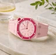 Pink silicone watch with round face displayed on white surface next to glass of water and green plant